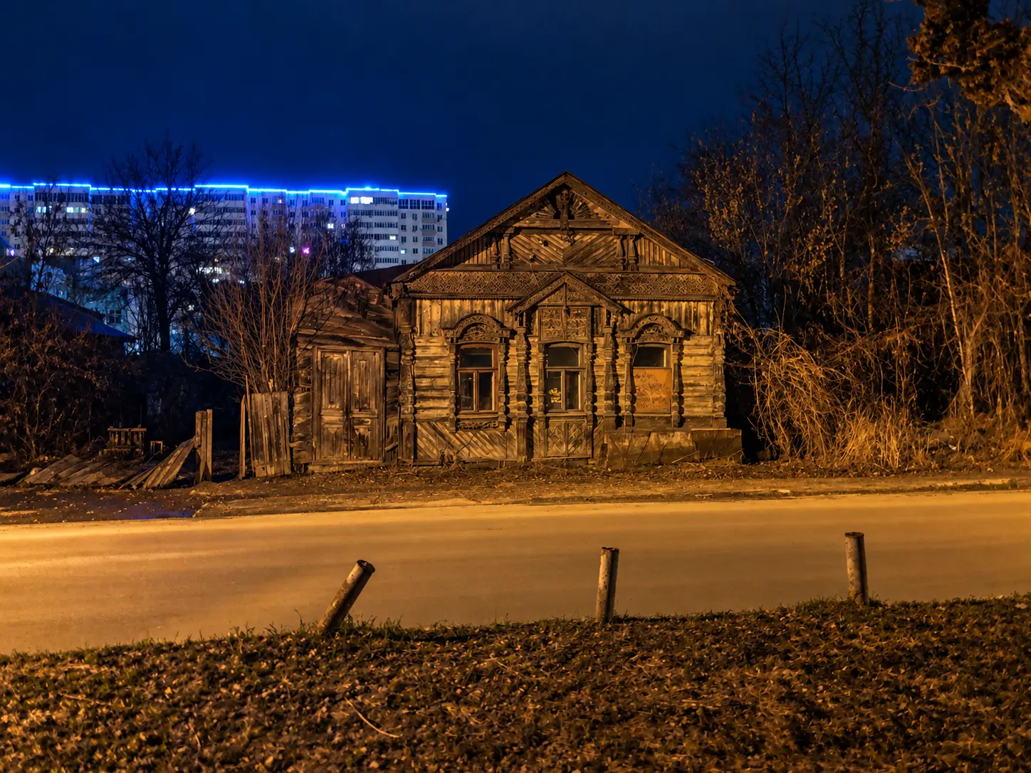 Старый деревянный дом ночью на фоне синей городской подсветки